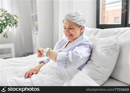 old age, technology and people concept - happy smiling senior woman in pajamas with smart watch sitting in bed at home bedroom. happy senior woman sitting in bed at home bedroom
