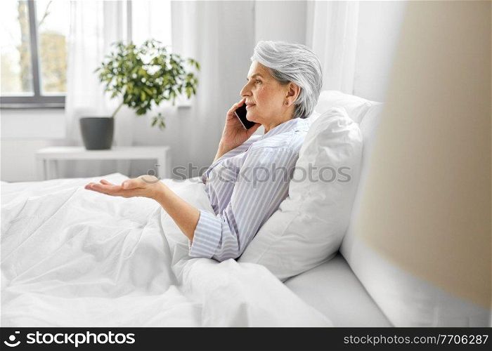 old age, technology and people concept - senior woman in pajamas calling on smartphone sitting in bed at home bedroom. senior woman calling on smartphone in bed at home