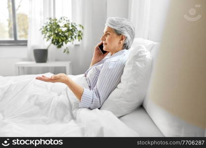 old age, technology and people concept - senior woman in pajamas calling on smartphone sitting in bed at home bedroom. senior woman calling on smartphone in bed at home