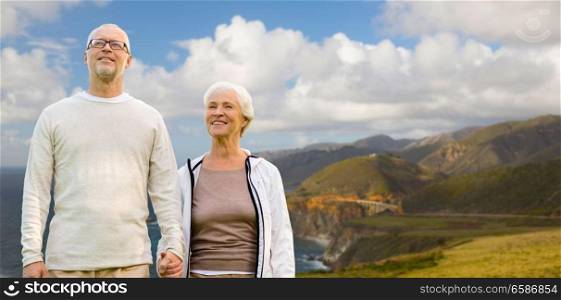 old age, tourism, travel and people concept - happy senior couple holding hands over bixby creek bridge on big sur coast of california background. happy senior couple over big sur coast