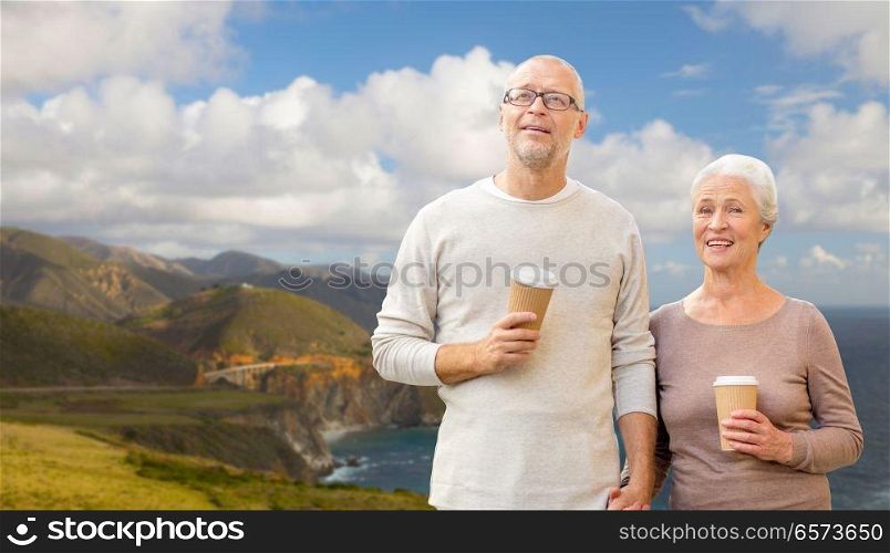 old age, tourism, travel and people concept - happy senior couple with coffee cups holding hands over bixby creek bridge on big sur coast of california background. senior couple with coffee cups over big sur coast