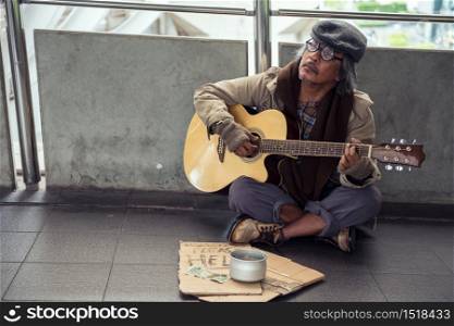 Old beggar or senior Homeless dirty man playing guitar flok song on footpath of modern city with donate bowl, dollar bill and coin, paper cardboard with help text to beg for money. Poverty in town