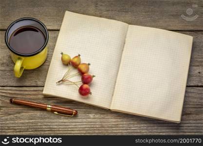 old blank notebook or travel journal with a metal mug of tea, pen and crab apples against rustic wood
