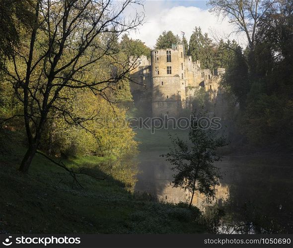 old castle of beaufort in luxemburg seen from side of pond on misty morning with tree silhouette