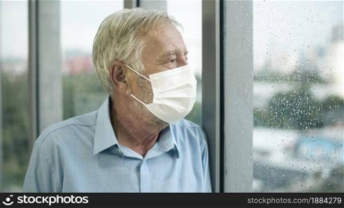 Old Caucasian man is wearing medical mask , looking to outside his home