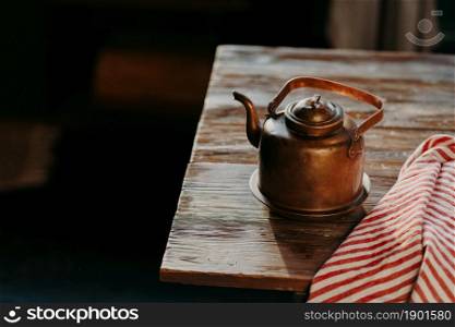 Old copper metal teapot on wooden table in dark room. Red striped towel nearby. Antique kettle for making tea or coffee. Cooking equipment