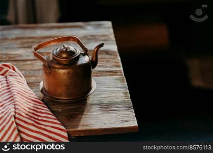 Old copper metal teapot on wooden table in dark room. Red striped towel nearby. Antique kettle for making tea or coffee. Cooking equipment