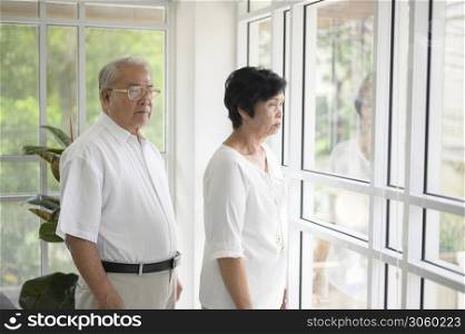 Old couple standing near the window looking outsideat home during quarantine due to COVID-19 pandemic
