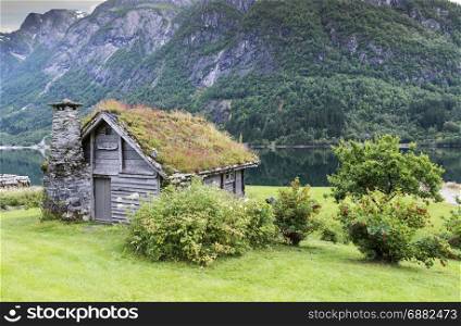 old fairytale-like house of wood with chimney of stacked slate and a roof with planting and trees on a fjord in Norway with garden with red flowers and green plants near Balastrand