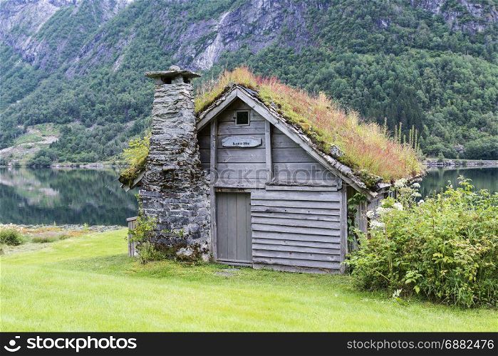 old fairytale-like house of wood with chimney of stacked slate and a roof with planting and trees on a fjord in Norway with garden with red flowers and green plants near Balestrand