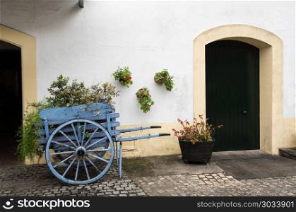 Old fashioned cart for horse with flowers by old white wall. Blue wooden horse cart filled with flowers and plants by old white painted stone building with arches. Old fashioned cart for horse with flowers by old white wall