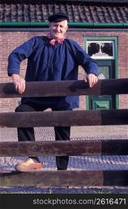 Old-fashioned senior man in traditional clothing and wooden clogs standing near fence