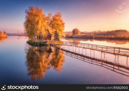 Old fisherman house and wooden pier at sunrise in autumn. Aerial view. Beautiful landscape with house on small island on the lake, colorful trees, jetty, reflection in water. Fall in Ukraine. Top view