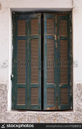 Old green wooden shutters are ajar on the old window against the background of the facade of the house.. Wooden peeled shutters on an old window.