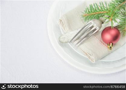 Old knife and fork, and red christmas ball and green spruce branche lie on the white porcelain plate, which is located on a table covered with a white tablecloth, top view