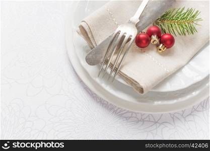 Old knife and fork, three red christmas balls and green spruce branche lie on the white porcelain plate, which is located on a table covered with a white tablecloth
