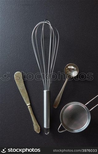 Old knife, whisk, spoon and sieve presented on a black concrete background with space for text. Top view. Set of kitchen tools knife, whisk, sieve and spoon on black concrete background with copy space. Top view