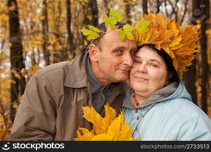 Old man and old woman with wreath of maple leaves embrace in autumnal forest