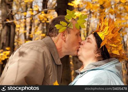 Old man and old woman with wreath of maple leaves kissing in autumnal forest