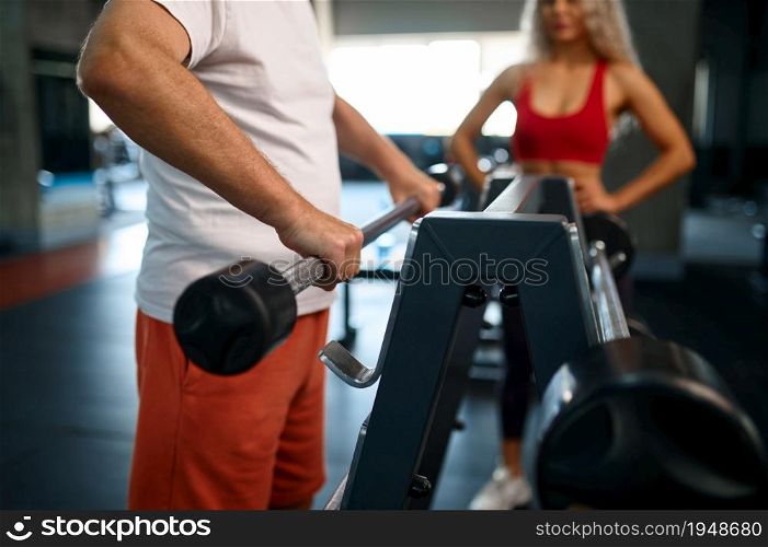 Old man doing exercise with bar, female personal trainer, gym interior on background. Sportive grandpa with woman instructor, workout. Old man doing exercise with bar, female trainer