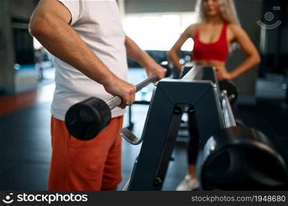 Old man doing exercise with bar, female personal trainer, gym interior on background. Sportive grandpa with woman instructor, workout. Old man doing exercise with bar, female trainer