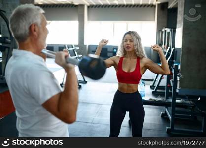 Old man doing exercise with bar, female personal trainer, gym interior on background. Sportive grandpa with woman instructor, workout in sport center. Old man doing exercise with bar, female trainer