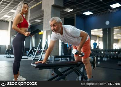 Old man doing exercise with dumbbell on bench, female personal trainer, gym interior on background. Sportive grandpa with woman instructor in sport center. Old man with dumbbell on bench, female trainer