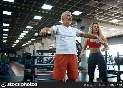Old man doing exercise with dumbbells, female personal trainer, gym interior on background. Sportive grandpa with woman instructor, training in sport center. Old man, exercise with dumbbells, female trainer