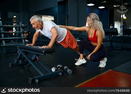 Old man doing push-up exercise on bench, female personal trainer, gym interior on background. Sportive grandpa with woman instructor in sport center. Old man, push-up exercise on bench, female trainer