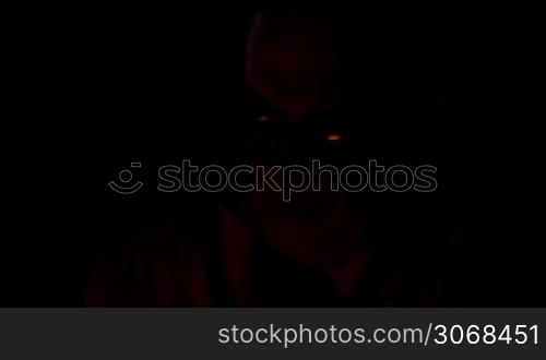 Old man in glasses sitting in the darkness with fire enlighting his face.