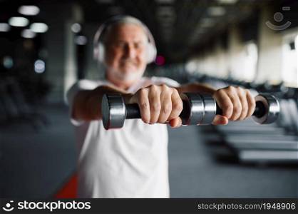 Old man in headphones doing exercise with dumbbells, treadmills and gym interior on background. Sportive grandpa on fitness training in sport center. Healty lifestyle, health care. Old man in headphones, exercise with dumbbells