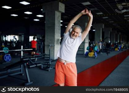 Old man in headphones doing fit exercise, gym interior on background. Sportive grandpa on fitness training in sport center. Healty lifestyle, health care. Old man in headphones doing fit exercise, gym