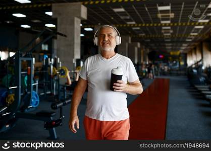 Old man in headphones drinks water, treadmills and gym interior on background. Sportive grandpa on fitness training in sport center. Healty lifestyle, health care. Old man in headphones drinks water in gym