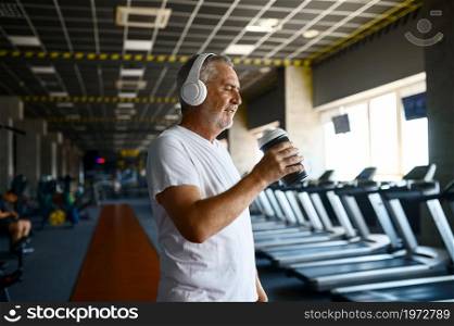 Old man in headphones drinks water, treadmills and gym interior on background. Sportive grandpa on fitness training in sport center. Healty lifestyle, health care. Old man in headphones drinks water in gym