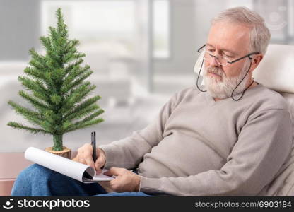 Old man making a shopping list with a pen on paper next to an undecorated fir tree for Christmas holidays