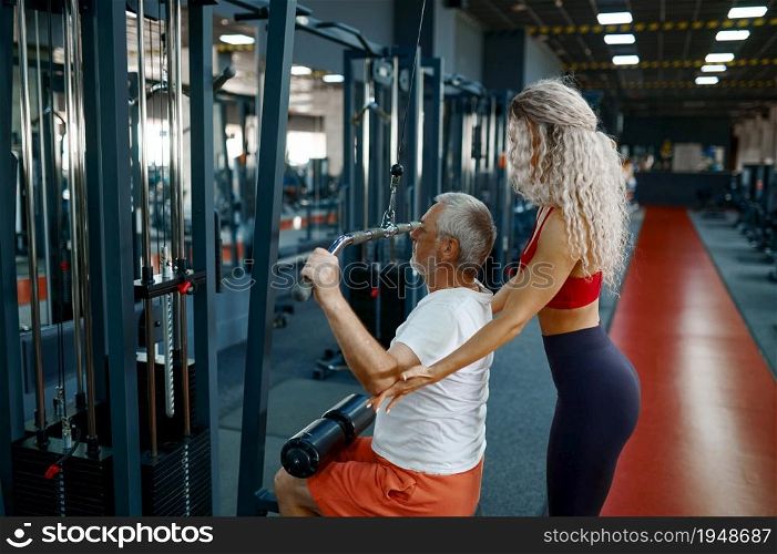 Old man on exercise machine, female personal trainer, gym interior on background. Sportive grandpa with woman instructor in sport center. Healty lifestyle, health care. Old man on exercise machine, female trainer, gym
