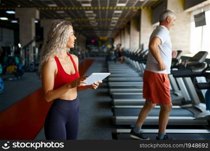 Old man on treadmill, female personal trainer with laptop, gym interior on background. Sportive grandpa with woman instructor, workout in sport center. Healty lifestyle, health care. Old man on treadmill, female trainer with laptop