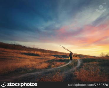 Old man silhouette walking a country road carrying dry tree branches on his shoulders, against sunset sky background. Idyllic rural landscape, late autumn season and dirt track across the golden hay.