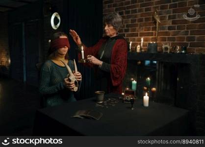 Old mature witch sorceress holding magic ritual for young female client. Wise woman blindfolding girl standing in atmospheric witchcraft office. Old mature witch sorceress holding magic ritual for young woman