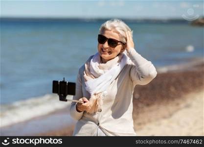 old people and leisure concept - happy smiling senior woman taking picture by smartphone and selfie stick on beach in estonia. senior woman taking selfie on beach
