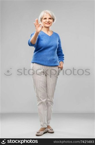 old people concept - portrait of smiling senior woman in blue sweater making ok gesture over grey background. portrait of smiling senior woman making ok gesture