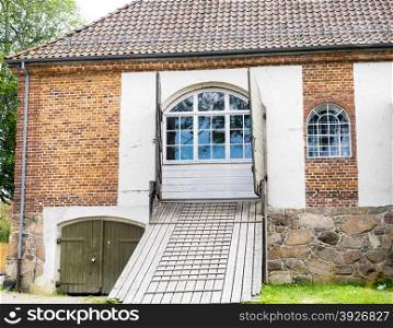 Old red brick building with wooden bridge path towards blue windows, dark roof stones
