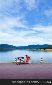 Old red motorcycle park by blue lake with summer sky and mountains in background. Nature scenery of Thailand
