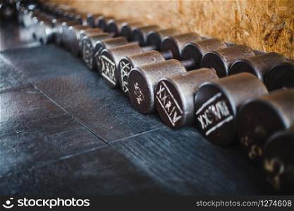 Old rusty dumbbells with roman numbers placed in a diagonal row on black resin covered gym floor.