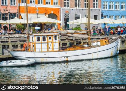 Old sailboat moored by Nyhavn promenade with walking tourists and restaurants, Copenhavn, Denmark