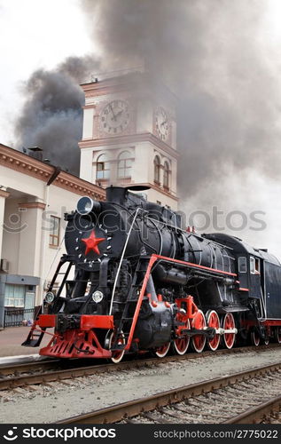 Old steam train is leaving a station. Vintage Steam engine locomotive train moving down railroad track towards camera.