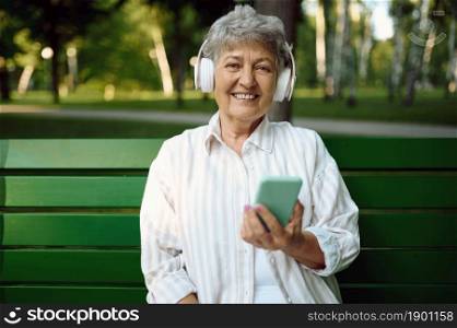 Old woman in headphones listens to music on the bench in summer park. Aged people lifestyle. Pretty grandmother having fun outdoors, an elderly female person on nature. Old woman in headphones listens to music on bench