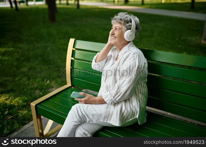 Old woman in headphones listens to music on the bench in summer park. Aged people lifestyle. Pretty grandmother having fun outdoors, an elderly female person on nature. Old woman in headphones listens to music on bench