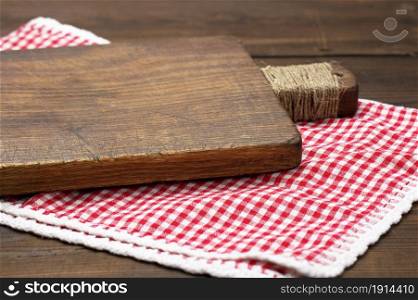 old wooden cutting board and folded red and white cotton kitchen napkin on a wooden brown background, top view, copy space