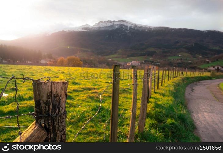 Old wooden pole of wire fence on blur green grass field, city in valley, and mountain in Europe. Fence of green animal grazing pasture beside the road. Wooden pole of green grass field with sunlight.